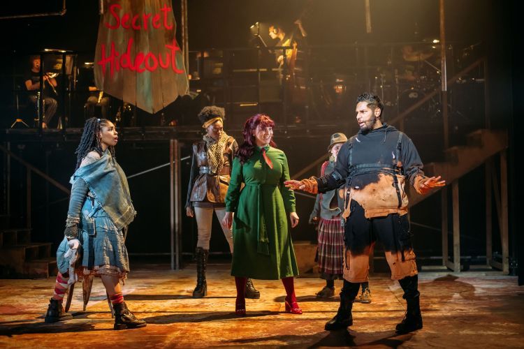 Actors in post-apocalyptic costumes on stage in Urinetown, with the band visible on a raised level in the background